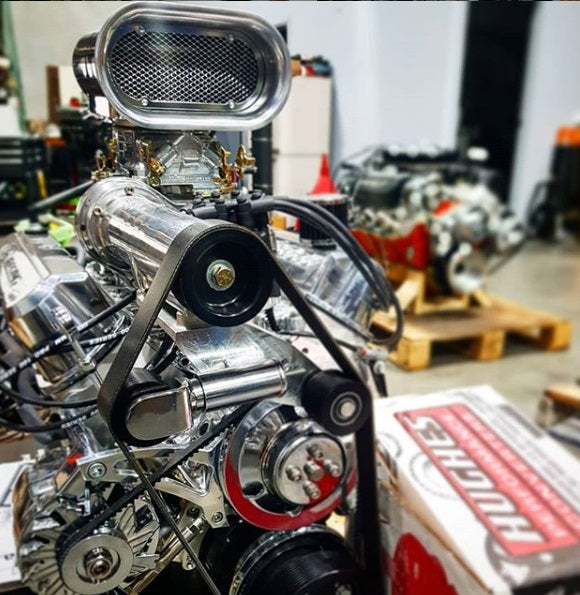 A highly polished chrome muscle engine with a large carburetor and pulley system, photographed in a workshop setting with a H&R box in the foreground and another engine on a pallet in the background.